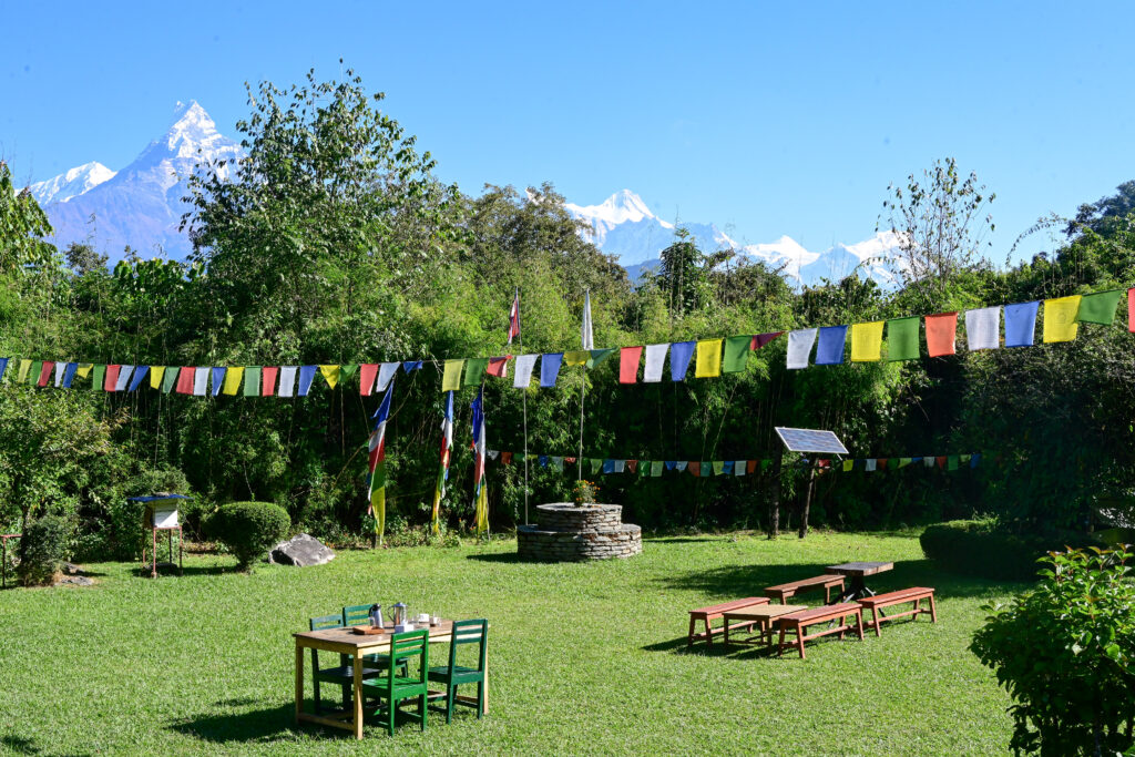 A sunny garden with colorful prayer flags strung across, green tables and benches, and snow-capped mountains in the distance.