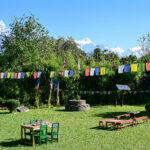 A sunny garden with colorful prayer flags strung across, green tables and benches, and snow-capped mountains in the distance.
